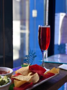 A glass of red drink on a table next to a plate of colorful tortilla chips with dips in small bowls.
