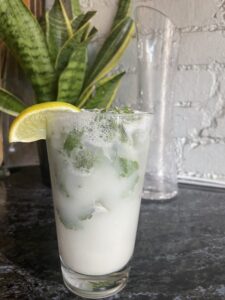 A frothy drink in a glass with ice, garnished with a lemon wedge and mint leaves, sits on a dark countertop with a tall plant and a clear glass pitcher in the background.