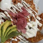 Plate of chilaquiles topped with avocado slices, pickled onions, crumbled cheese, and drizzled with sour cream and a dollop of cream.