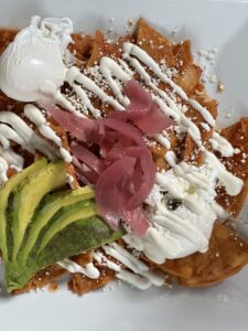 Plate of chilaquiles topped with avocado slices, pickled onions, crumbled cheese, and drizzled with sour cream and a dollop of cream.