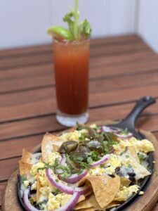 A plate of nachos topped with cheese, scrambled eggs, red onions, jalapenos, and cilantro is on a wooden table with a tall beverage garnished with celery and lime in the background.
