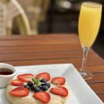 A pancake topped with strawberries, blueberries, and a mint leaf, served on a white plate with a side of syrup and a glass of orange juice on a wooden table.