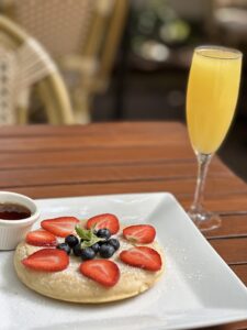 A pancake topped with strawberries, blueberries, and a mint leaf, served on a white plate with a side of syrup and a glass of orange juice on a wooden table.
