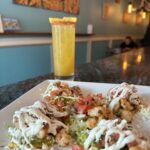 A plate of shrimp tacos drizzled with a white sauce is on a bar counter with a tall yellow drink garnished with an orange slice in the background.