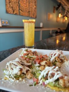 A plate of shrimp tacos drizzled with a white sauce is on a bar counter with a tall yellow drink garnished with an orange slice in the background.