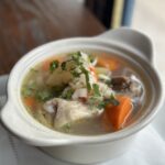 A white ceramic bowl filled with a clear soup containing pieces of chicken, carrots, and other vegetables, garnished with chopped cilantro, sits on a napkin with two lime wedges beside it.