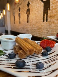 A plate of churros with two dipping sauces, garnished with blueberries, strawberries, and a drizzle of chocolate sauce, set on a table in a restaurant with a mural on a brick wall in the background.