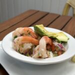 A bowl of shrimp ceviche with diced tomatoes, red onions, herbs, and slices of avocado, served on a wooden table.