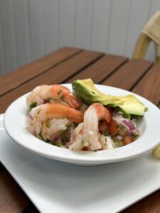 A bowl of shrimp ceviche with diced tomatoes, red onions, herbs, and slices of avocado, served on a wooden table.