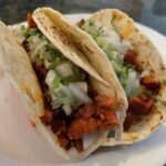 Two tacos on a white plate, each filled with chopped seasoned meat, diced onions, and cilantro.