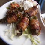 Plate of bacon-wrapped jalapeño poppers with a side of diced vegetables, sliced radishes, and a dollop of sour cream on shredded lettuce.