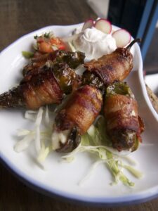 Plate of bacon-wrapped jalapeño poppers with a side of diced vegetables, sliced radishes, and a dollop of sour cream on shredded lettuce.