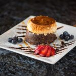 A dessert with a layered cake topped with caramel on a white plate, garnished with strawberry slices, blueberries, and chocolate drizzle.