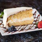 A slice of layered vanilla cake with white frosting on a plate garnished with blueberries, a strawberry, and a mint leaf, drizzled with chocolate sauce.