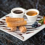 A plate with three churros dusted with powdered sugar, accompanied by chocolate and caramel dipping sauces, strawberries, and blueberries.