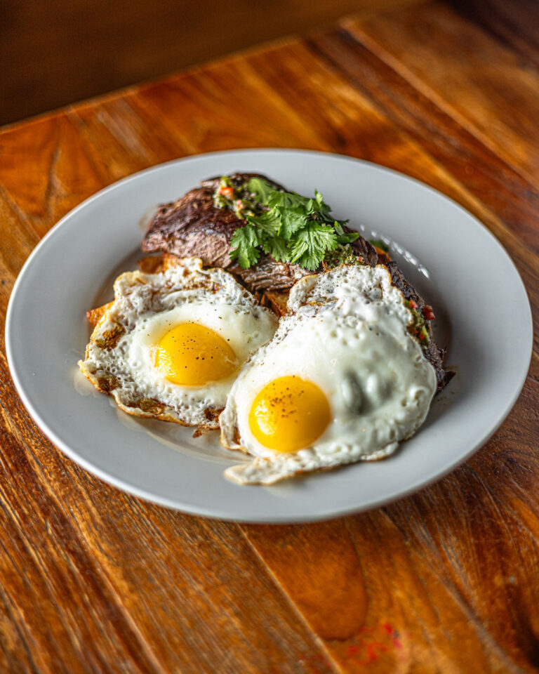 A plate with two sunny-side-up eggs and a slice of steak garnished with cilantro sits on a wooden table.