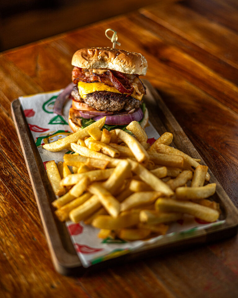 A burger with bacon and cheese on a sesame seed bun served with a side of fries on a wooden tray.