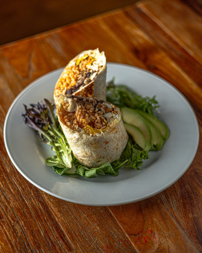 A burrito cut in half is served on a white plate, placed on a bed of lettuce and greens, with avocado slices on the side.