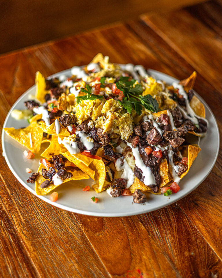 A plate of nachos topped with cheese, ground meat, black beans, chopped tomatoes, sour cream, and herbs on a wooden table.