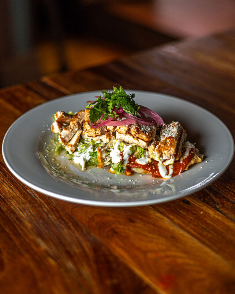 A plated dish featuring sliced meat on a bed of greens, topped with pickled onions and herbs, drizzled with sauce.