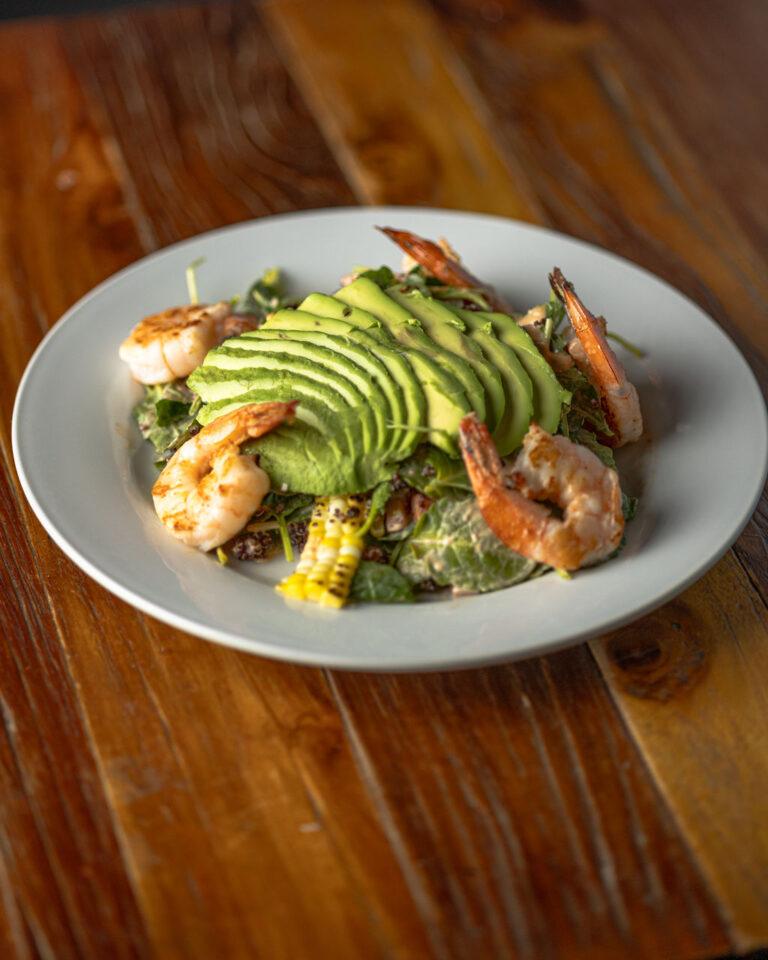 A plate of salad topped with sliced avocado and grilled shrimp on a wooden table.